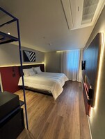 Quarto de hotel moderno com beliche, escrivaninha e uma janela grande com cortinas.