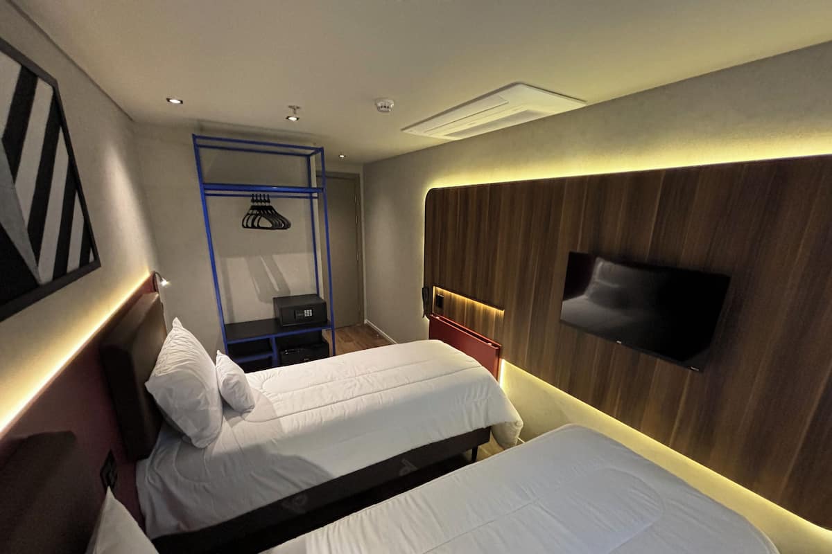 Une chambre d’hôtel avec deux lits, un téléviseur fixé au mur, un bureau avec une chaise et une petite étagère avec une lampe.