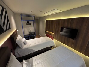 Een hotelkamer met twee bedden, een aan de muur bevestigde televisie, een bureau met stoel, en een klein plankje met een lamp.