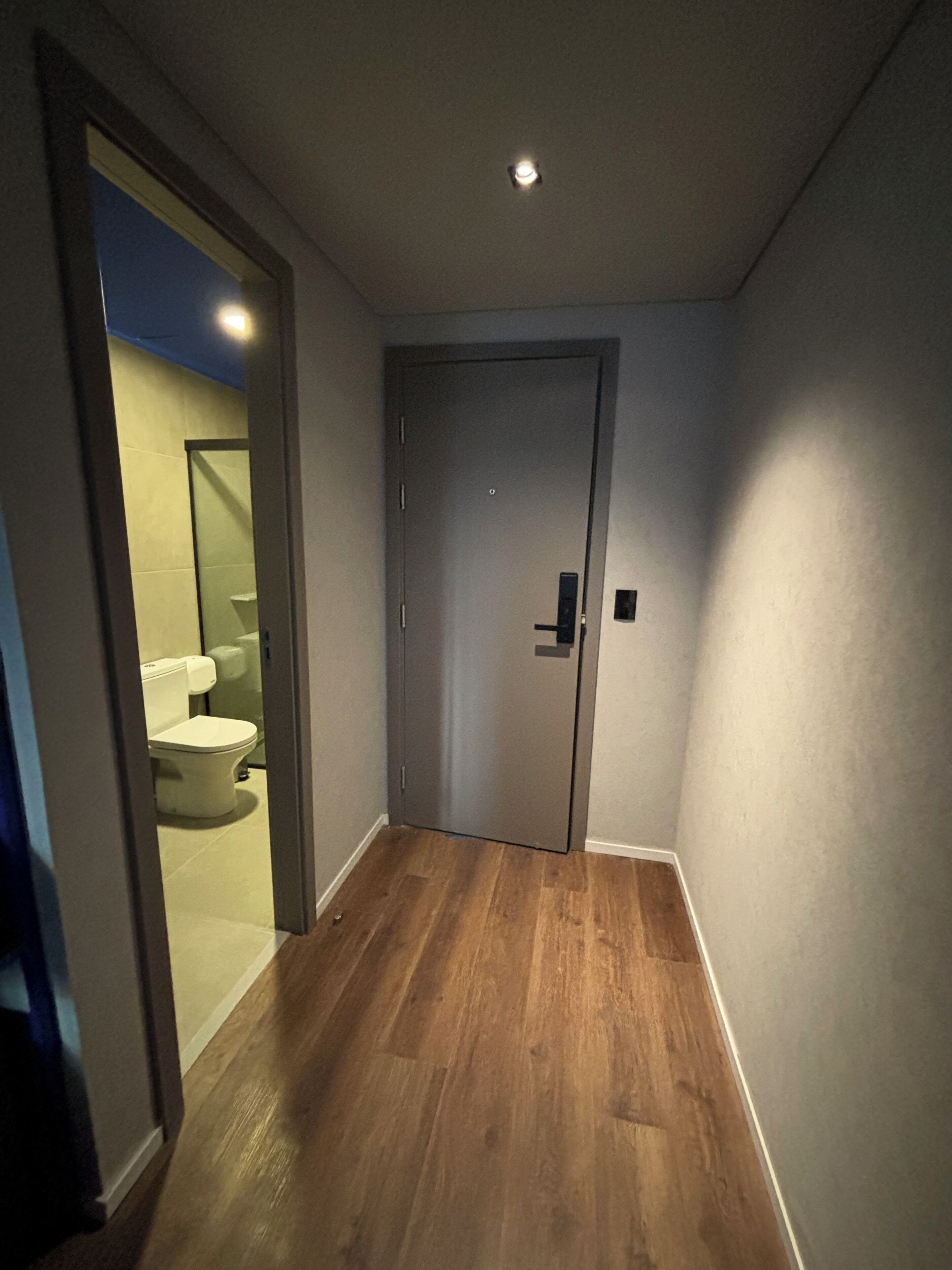 Un pasillo estrecho con piso de madera, una puerta cerrada y un inodoro a la vista en la habitación contigua.
