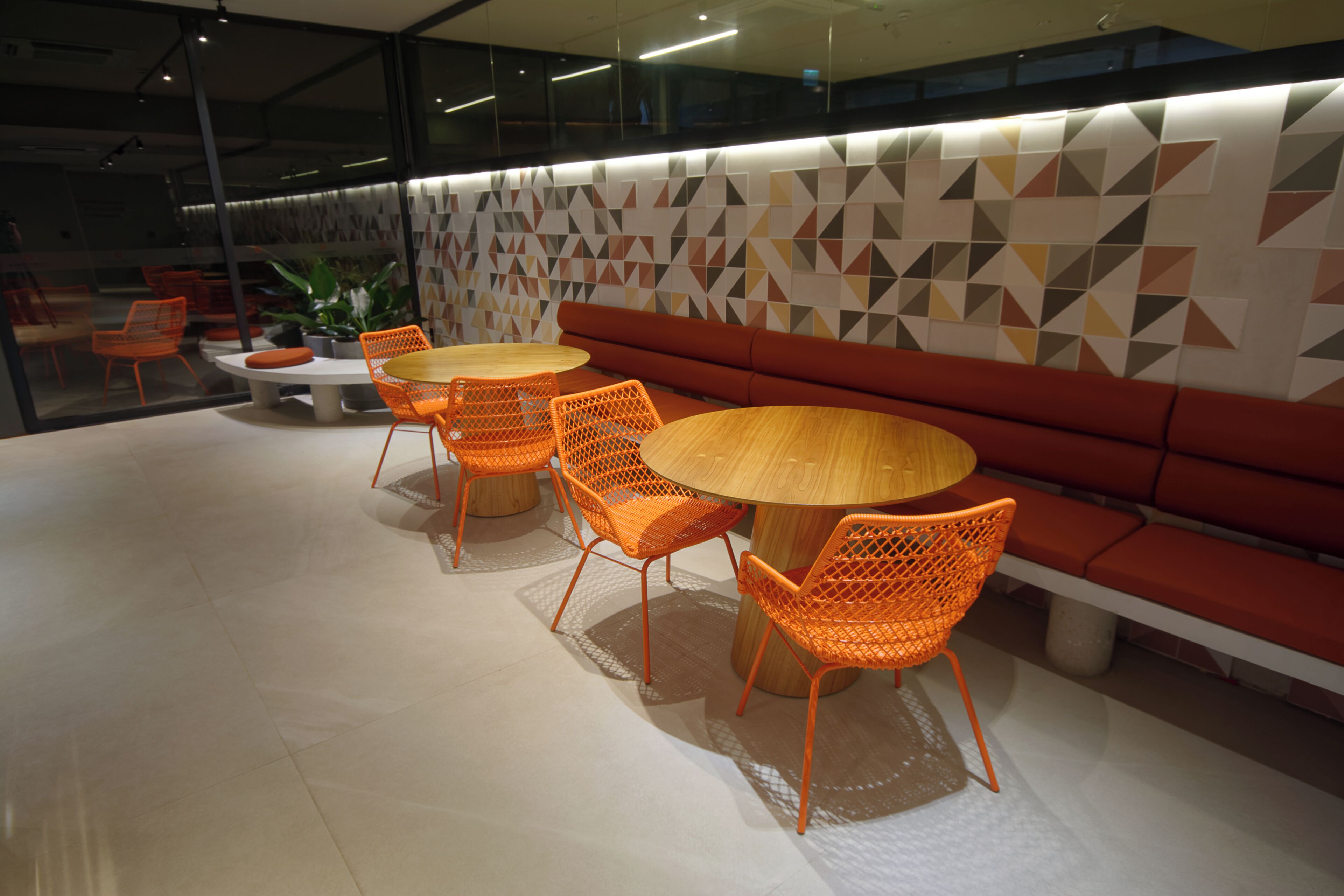 Un área de descanso moderna con una mesa redonda de madera y sillas color naranja, un sofá rojo y una pared con estampado geométrico.