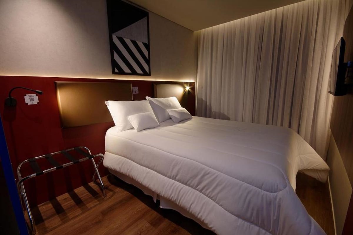 Une chambre d’hôtel dotée d’un grand lit, d’une tête de lit avec éclairage intégré, d’un téléviseur fixé au mur et d’une fenêtre avec des rideaux.
