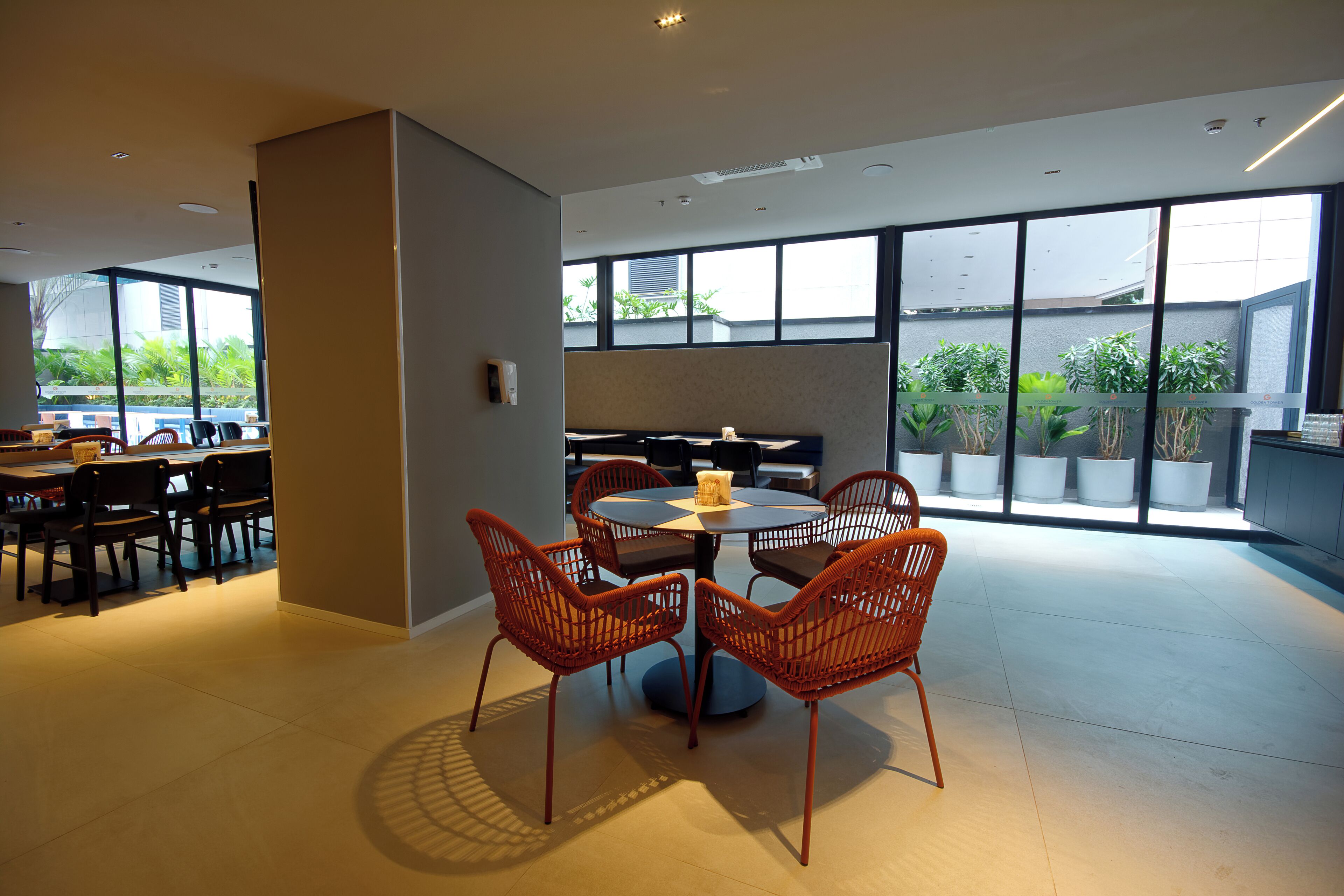 Un comedor moderno con una mesa redonda y cuatro sillas de mimbre, ventanas amplias con vistas a la vegetación y una zona de cocina al fondo.