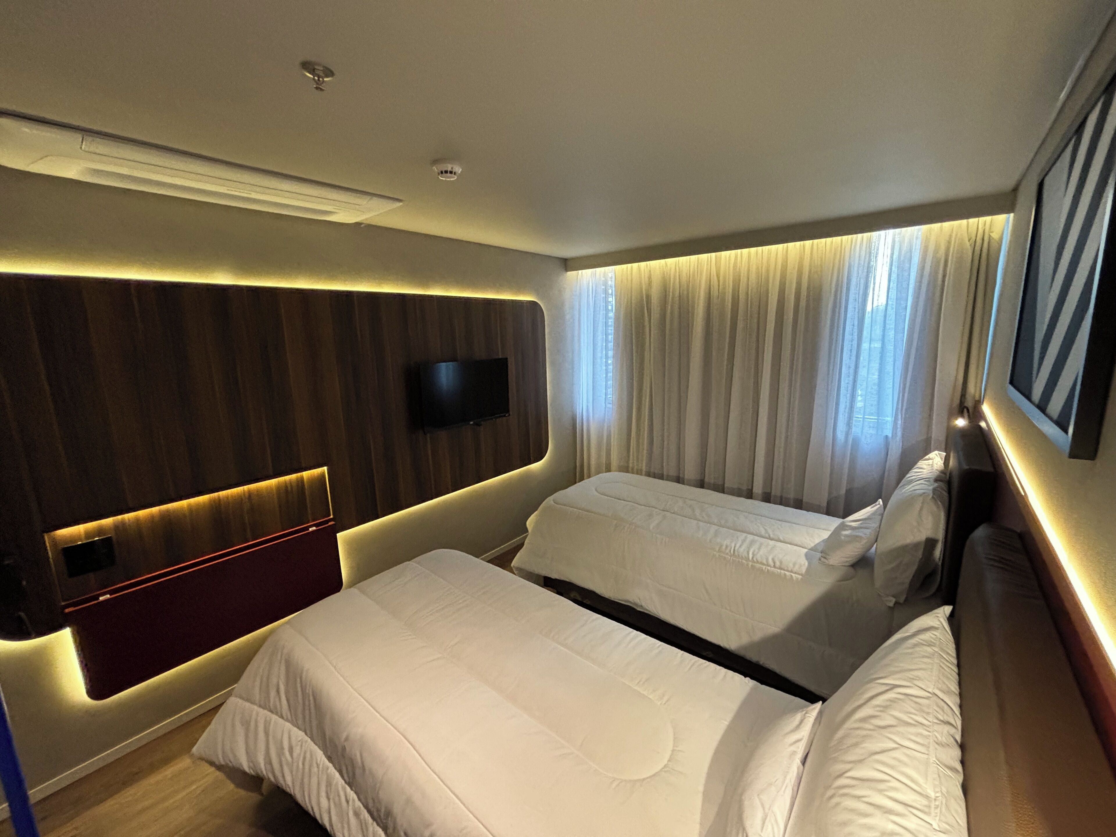 A hotel room with two beds, a TV mounted on the wall, and a window with curtains.