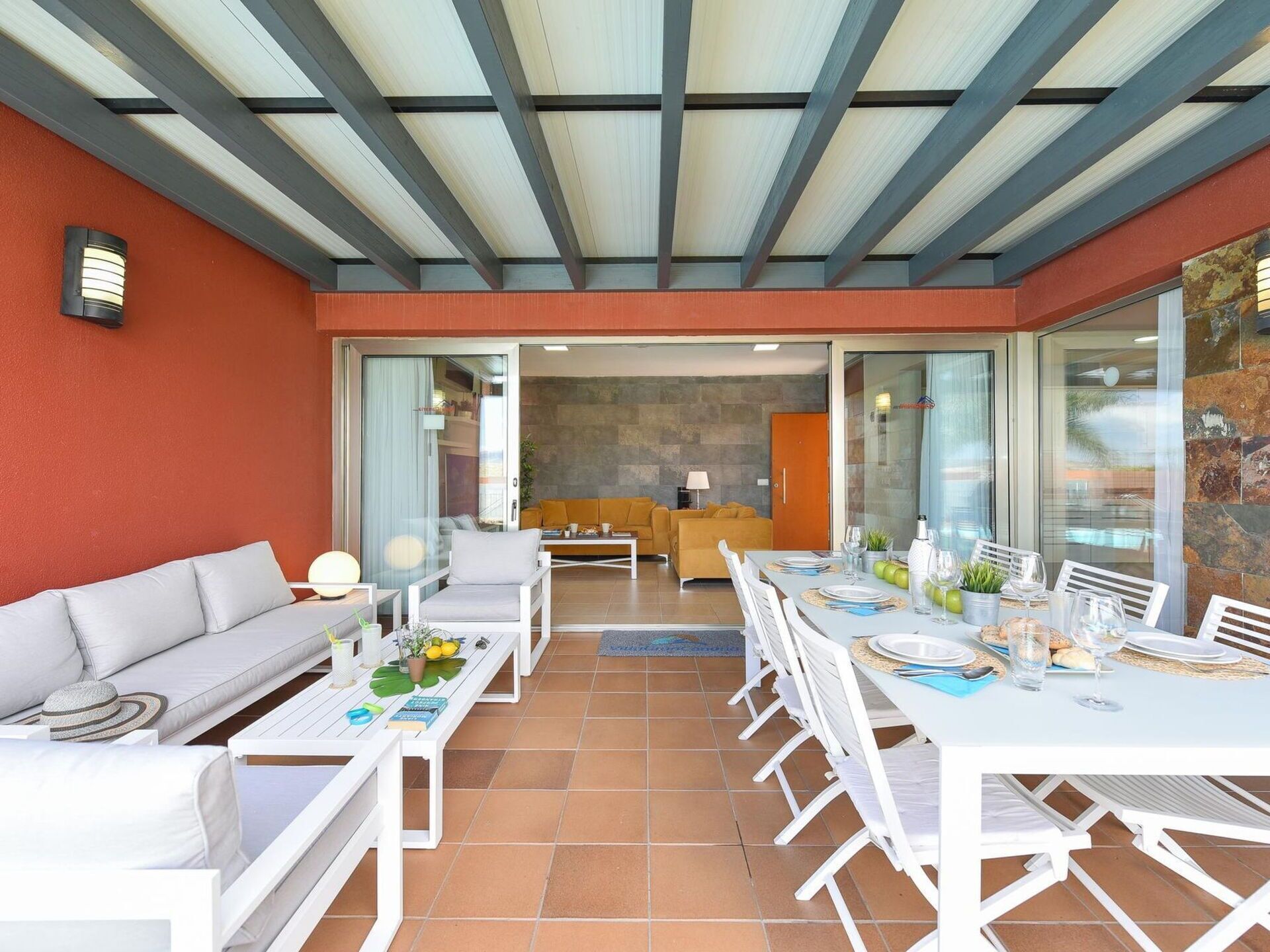 Un área de comedor exterior moderna con mobiliario blanco, un muro de piedra y vistas del interior.