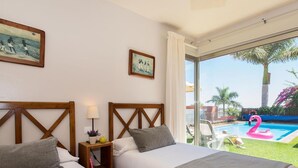 Ein Hotelzimmer mit zwei Betten, einem Nachttisch, einer Lampe und einem Fenster mit Blick auf einen Swimmingpool und Palmen.