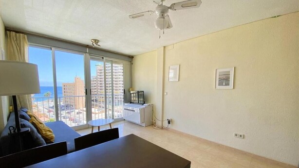 A room with a dining table, chairs, a sofa, a ceiling fan, a window with a view of buildings, and a small table with a lamp. - Apartamento Miramar Calpe (Calpe)