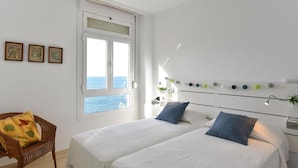 A bedroom with two beds, a window with a view of the sea, a wicker chair, and a wall with framed pictures. - Nice apartment for 4 guests with WIFI, TV, terrace and pets allowed (Las Palmas De Gran Canaria)