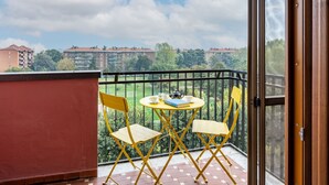 Un balcón con mesa y sillas, vistas a edificios y follaje, y una puerta a la derecha.