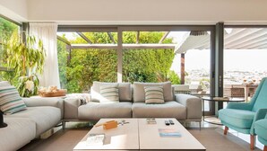 Ein modernes Wohnzimmer mit einer Eckcouch, einem Couchtisch und einem Türkis-farbenen Sessel. Ein großes Fenster bietet Blick auf Grünflächen und ein Gebäude in der Ferne.
