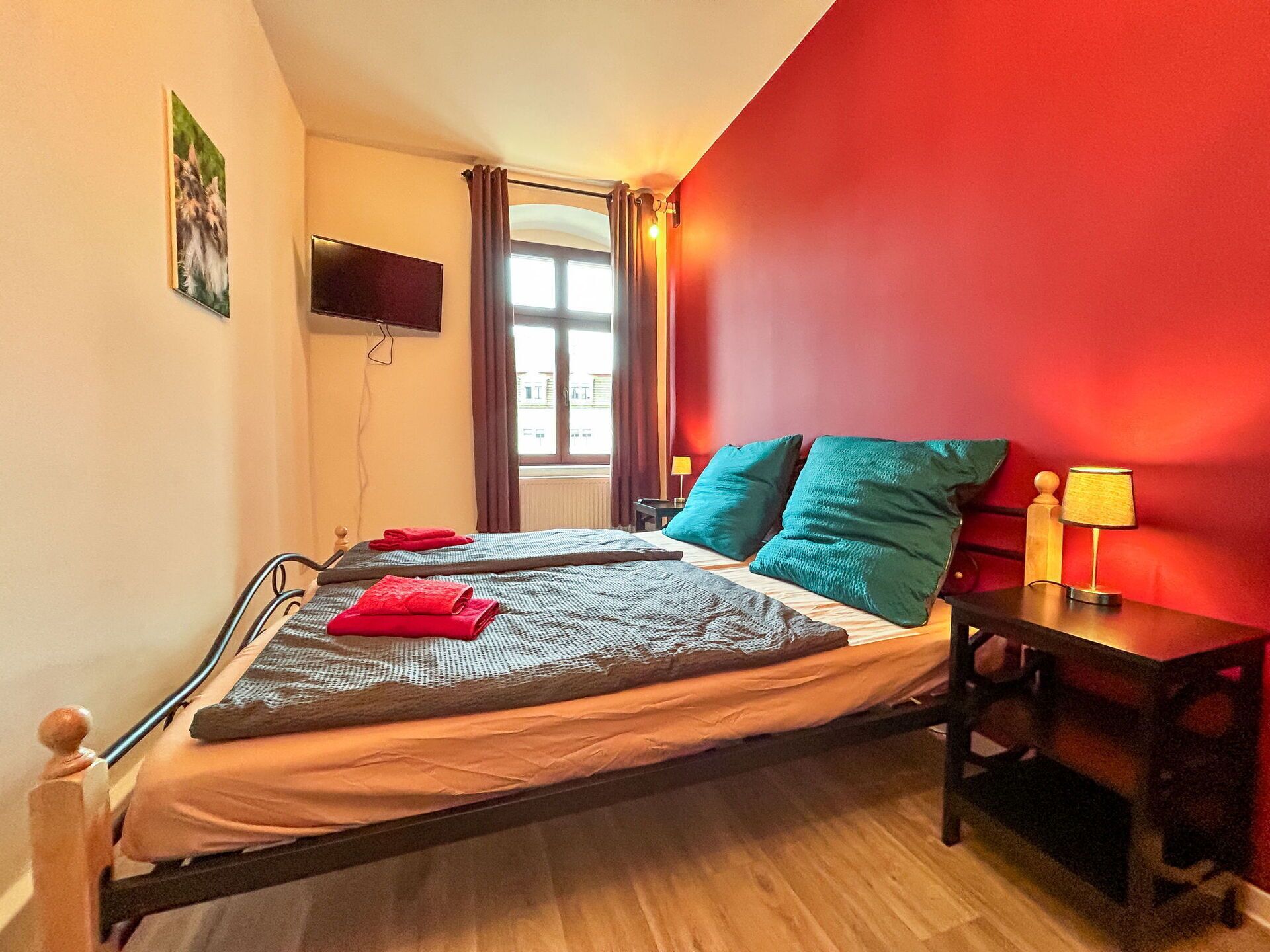A single bed with a grey blanket and red towels, a wooden nightstand with a lamp, a TV on the wall, and a window with curtains.