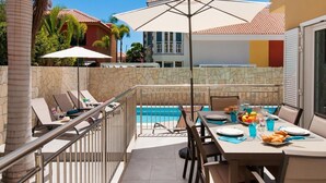 Un espace repas aménagé au bord de la piscine, avec une table dressée pour deux personnes, une piscine et une vue sur un bâtiment doté d’un toit en tuiles.