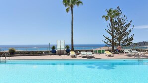 Une piscine d’un bleu éclatant, bordée de chaises longues et de palmiers, avec vue sur une plage et l’océan.