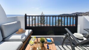 Un balcon avec vue sur la mer, un canapé, une table avec des boissons et un livre, et une chaise.