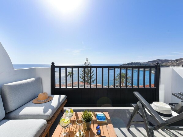 A balcony with a view of the sea, a sofa, a table with drinks and a book, and a chair.