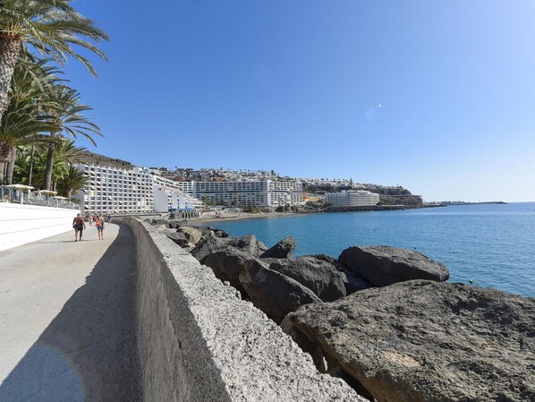 A coastal promenade with a rocky breakwater, palm trees, and a row of white hotels with multiple floors and balconies overlooking the sea.