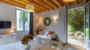 Une chambre moderne avec un plafond en bois, un lit recouvert de linge de lit blanc, un téléviseur posé sur un meuble blanc et une table à manger agrémentée de verres à vin et d’un vase de fleurs.