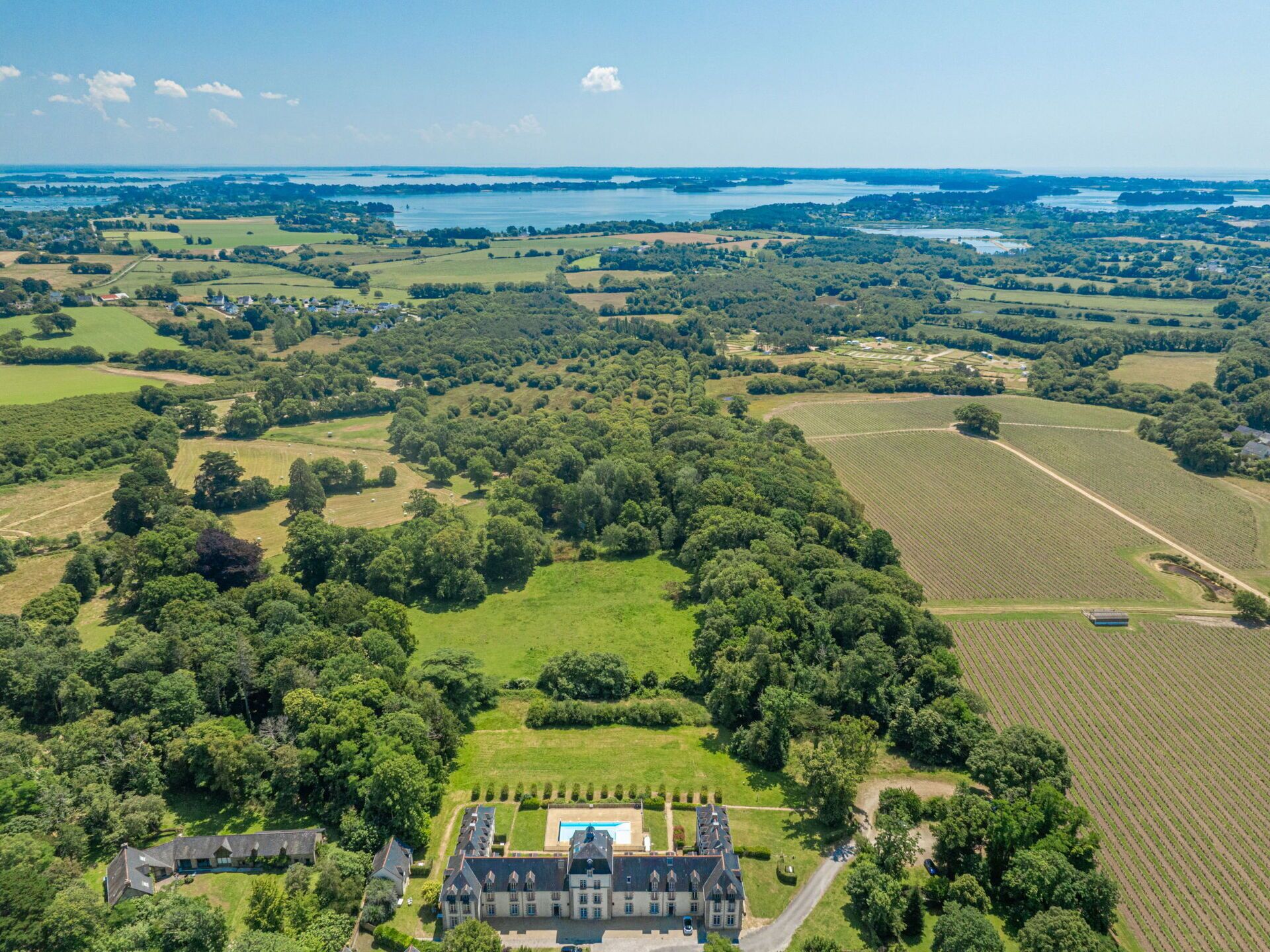 Une vue aérienne d’un vaste domaine, avec de larges champs verdoyants, un bâtiment central doté d’une piscine et une rivière en arrière-plan.