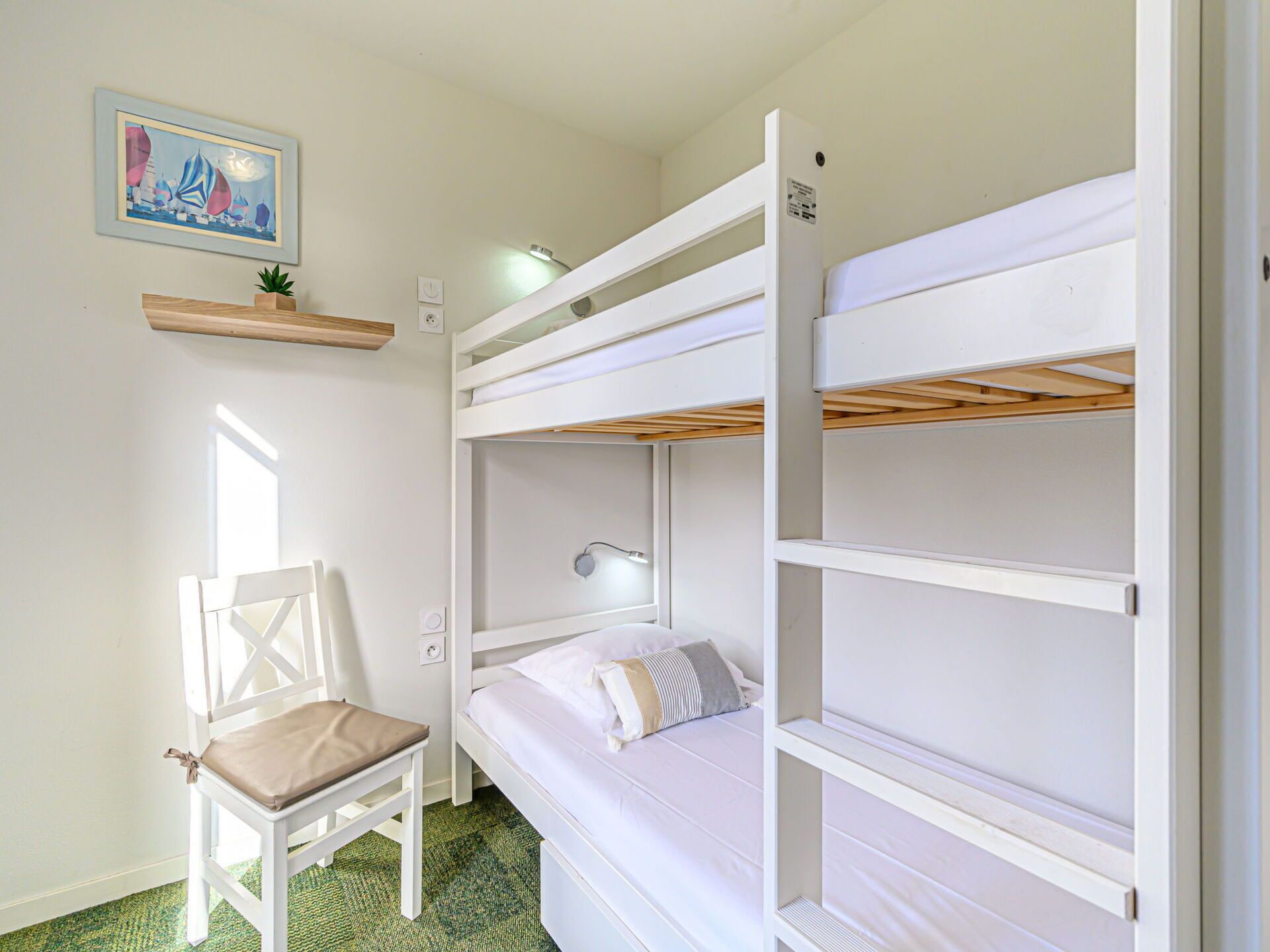 A bunk bed room with a chair, a small shelf with a plant, and a painting on the wall.