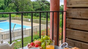 Ett frukostbord med croissanter, frukt, juice och en tekanna på en balkong med utsikt över en pool och en trädgård.