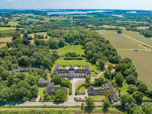 Une vue aérienne d’un vaste domaine comprenant plusieurs bâtiments, de vastes champs verdoyants et une étendue d’eau en arrière-plan.