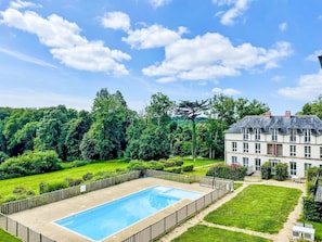 A large rectangular swimming pool surrounded by a wooden fence, with a well-maintained lawn and a large building with multiple windows and a balcony in the background.