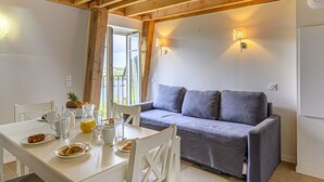 Ein gemütlicher Raum mit Holzbalkendecke, einem grauen Sofa, einem Esstisch mit Frühstücksgebäck und Blick durch eine Tür.