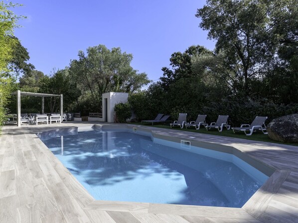 En rektangulär swimmingpool omgiven av ett trädäck, en pergola med sittplatser och solstolar längs poolkanten.