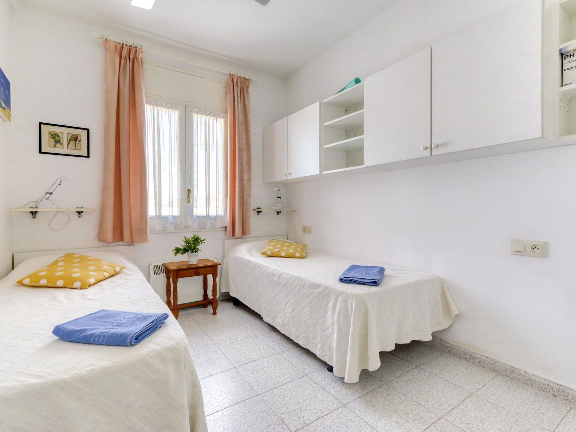 Une petite chambre propre, avec deux lits, une petite table, une plante et une fenêtre avec des rideaux.