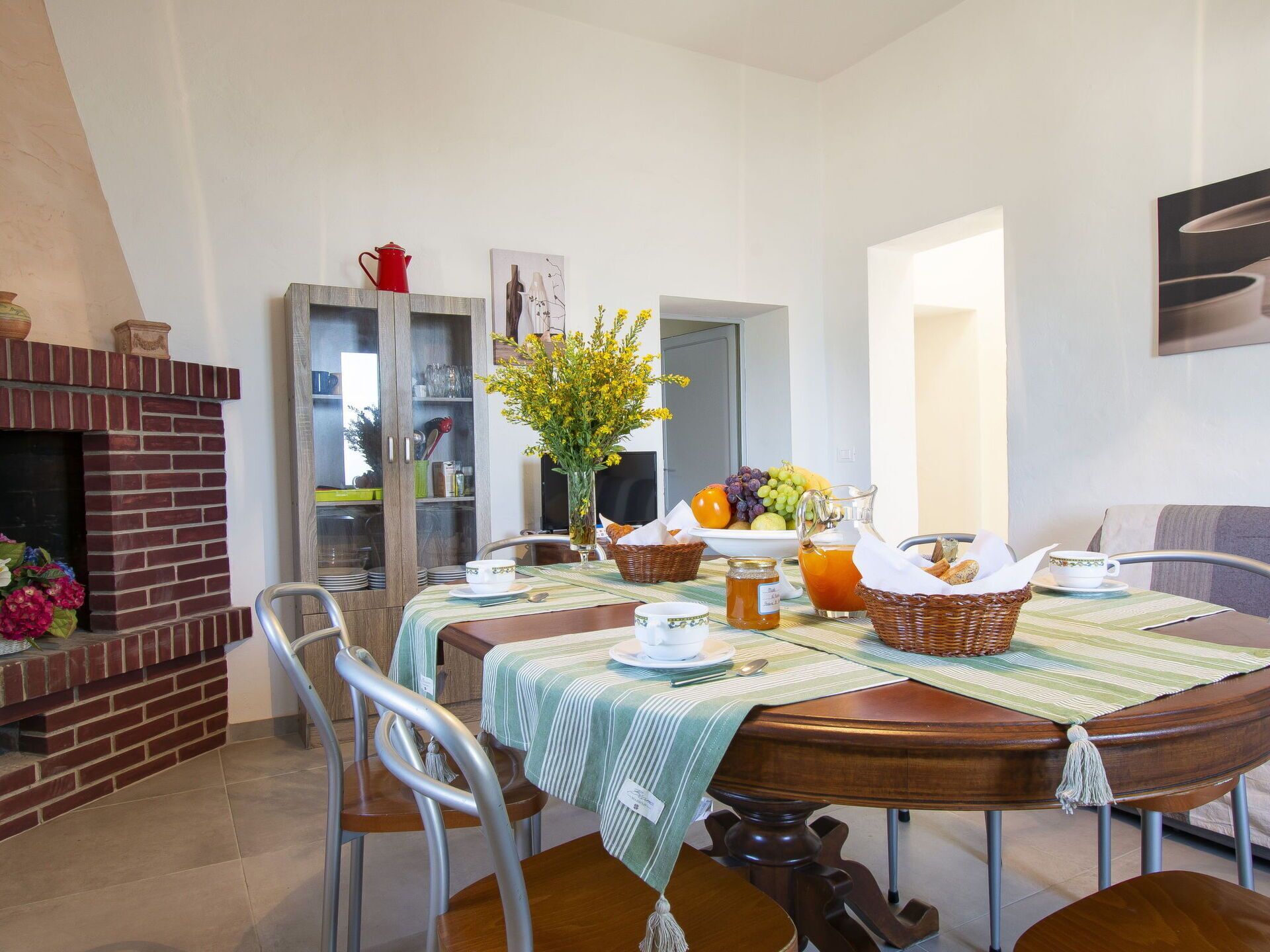 A dining area with a brick fireplace, a dining table set with plates, a vase of flowers, and a fruit bowl.
