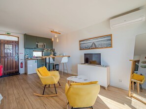 Un espacio habitable compacto con kitchenette, una zona de comedor con sillas amarillas, un televisor y una pintura de paisaje marino.
