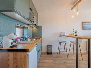 Cocina moderna con encimera de madera, lavavajillas integrado y un lavabo. Se observa una zona de comedor con una mesa y sillas, y un cubo de basura.