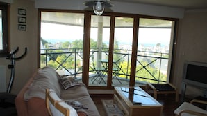 A living room with a sofa, a table, and a TV. There is a balcony with a view of the city. - Douarnenez, Proche de la Plage des Sables Blancs à Tréboul (Douarnenez)