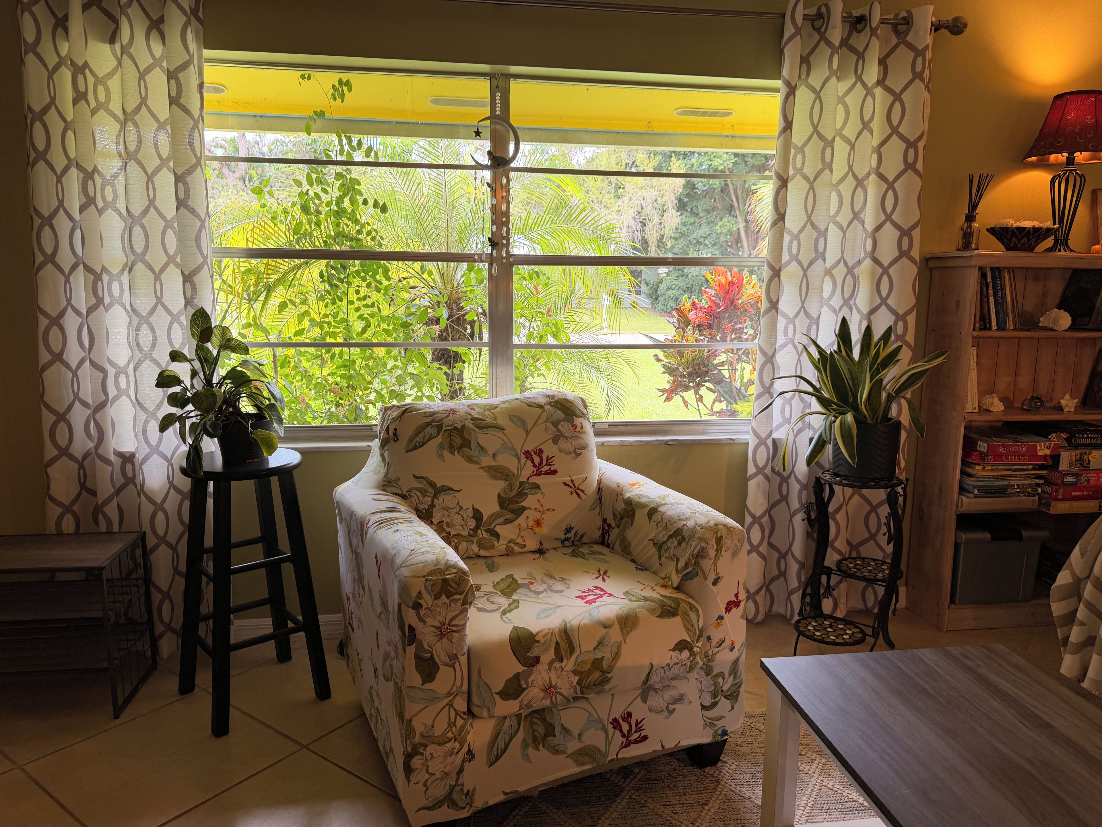Une pièce comprenant un fauteuil à motifs floraux, une petite table, une bibliothèque et une fenêtre donnant sur un paysage verdoyant.