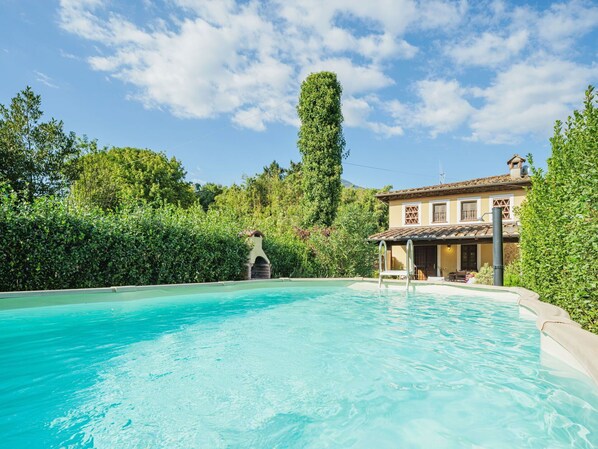 Relax by the serene pool surrounded by a lush hedge, adjacent to a charming villa with a tiled roof and chimney.