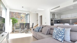 Una sala de estar moderna con un sofá gris, un comedor con mesa de vidrio y una cocina con gabinetes blancos y estufa gris.