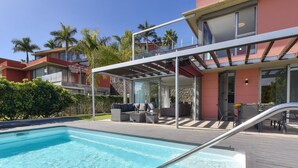 Un área de piscina moderna con una piscina transparente, una escalera de metal y mobiliario exterior.