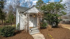 A small, single-story house with a front porch, surrounded by a garden with various plants and flowers. - Updated 1BR cottage on private acre+ lot (Winston-Salem)
