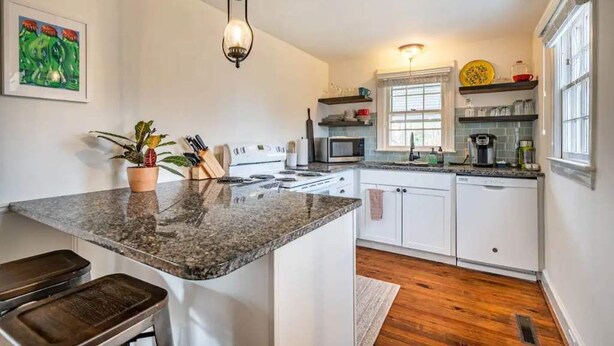 A kitchen with white cabinets, a granite countertop, a stove, and a microwave. There is a hanging light fixture and a window with blinds. - Updated 1BR cottage on private acre+ lot (Winston-Salem)