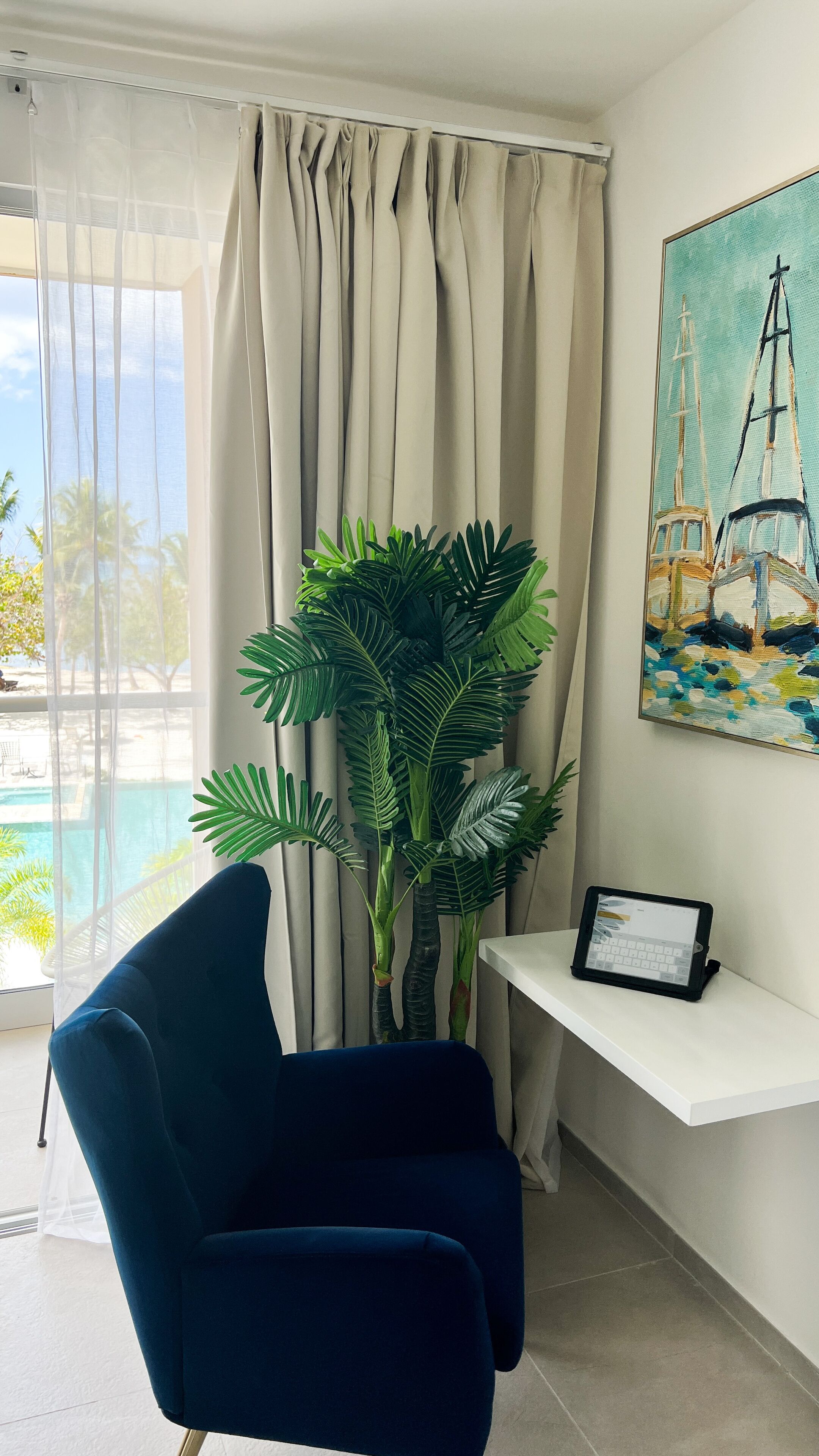 A room with a blue chair, a potted plant, a painting of sailboats, and a tablet on a white shelf.