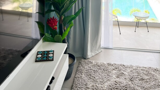 A modern living room with a large window, a potted plant, a small table, and a view of a pool and palm trees outside.