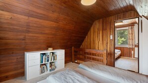 Une chambre confortable aux murs en bois, avec une commode blanche où sont posés des livres, un lit et une fenêtre agrémentée de rideaux.