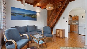 Un salon chaleureux avec un plafond en bois, un canapé, deux fauteuils, une table basse et une vue sur la cuisine et l’escalier.