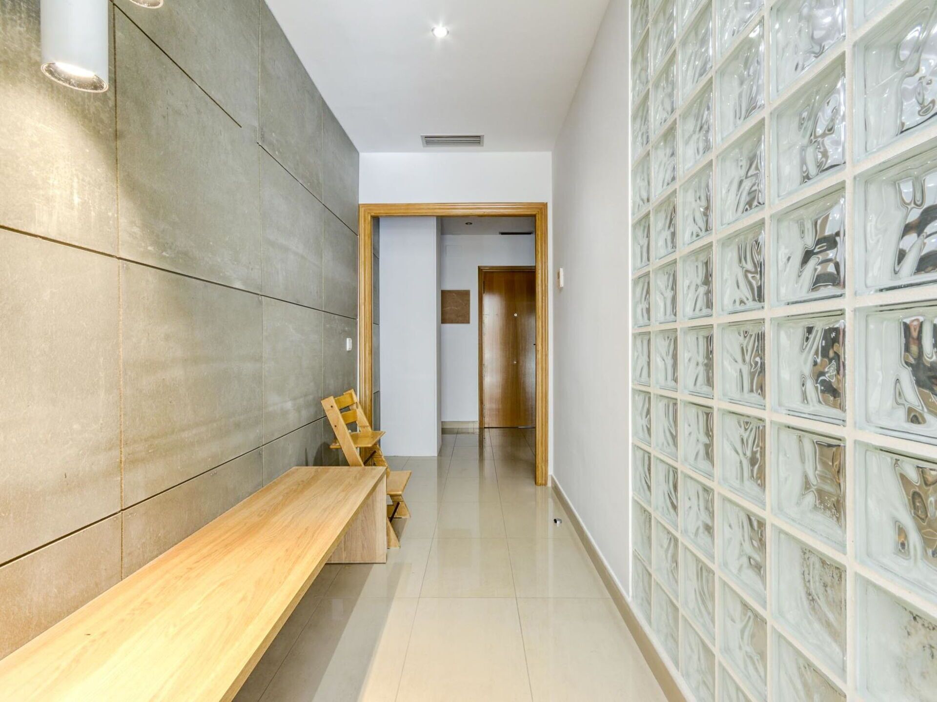 Un couloir avec un banc en bois, des murs carrelés et un mur de verre orné de motifs abstraits.
