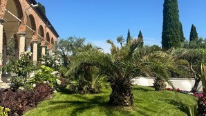 A courtyard with a brick building, palm trees, and a tall cypress tree. - Locanda al Santuario (Peschiera del Garda)
