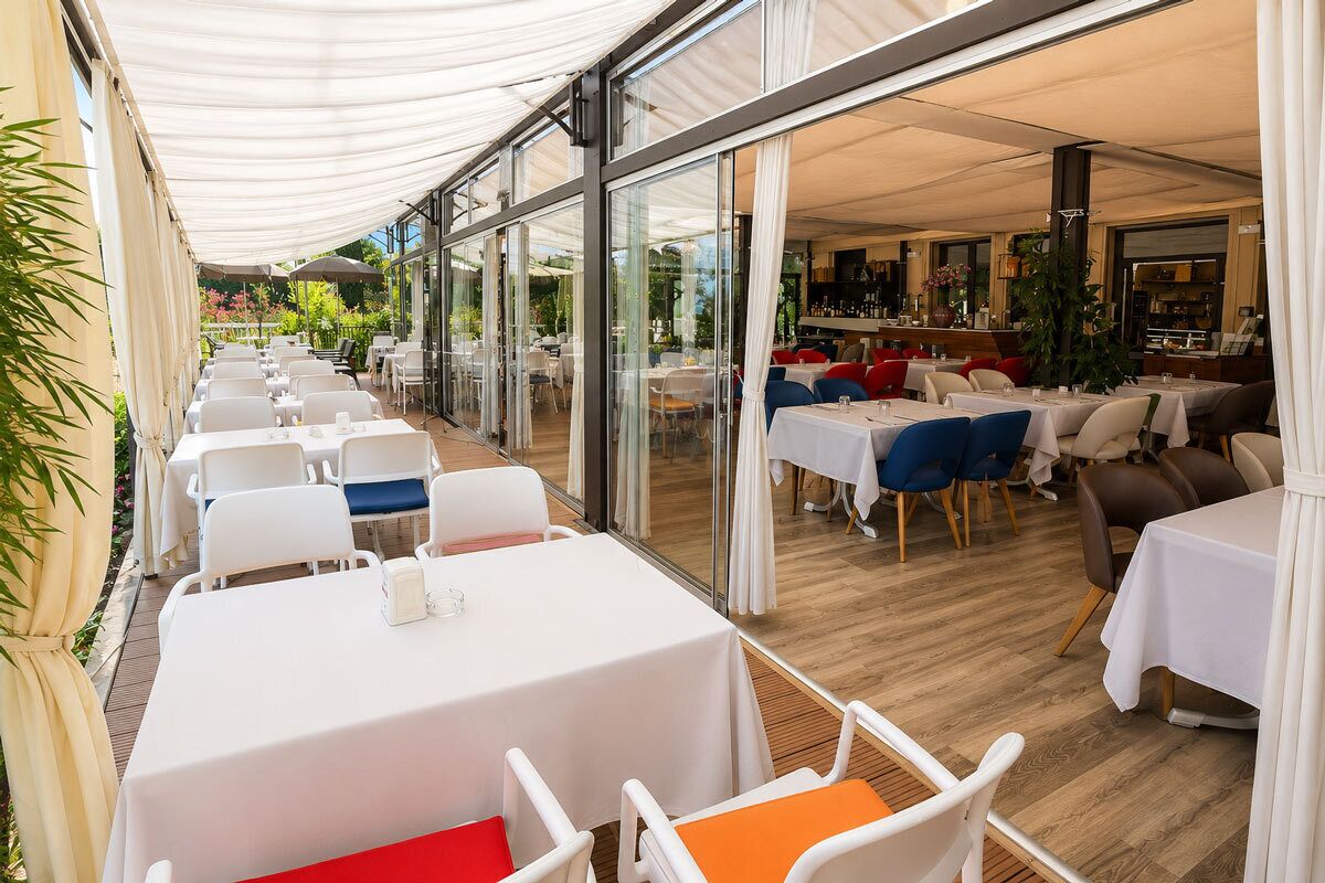 An outdoor dining area with tables and chairs, a glass wall, and a view of the garden.