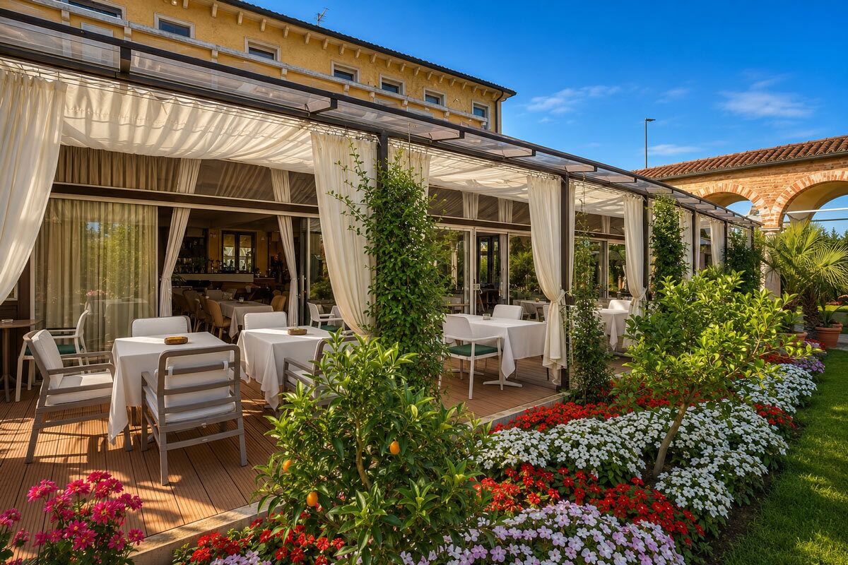 An outdoor dining area with tables and chairs, a covered patio, and a garden with colorful flowers.
