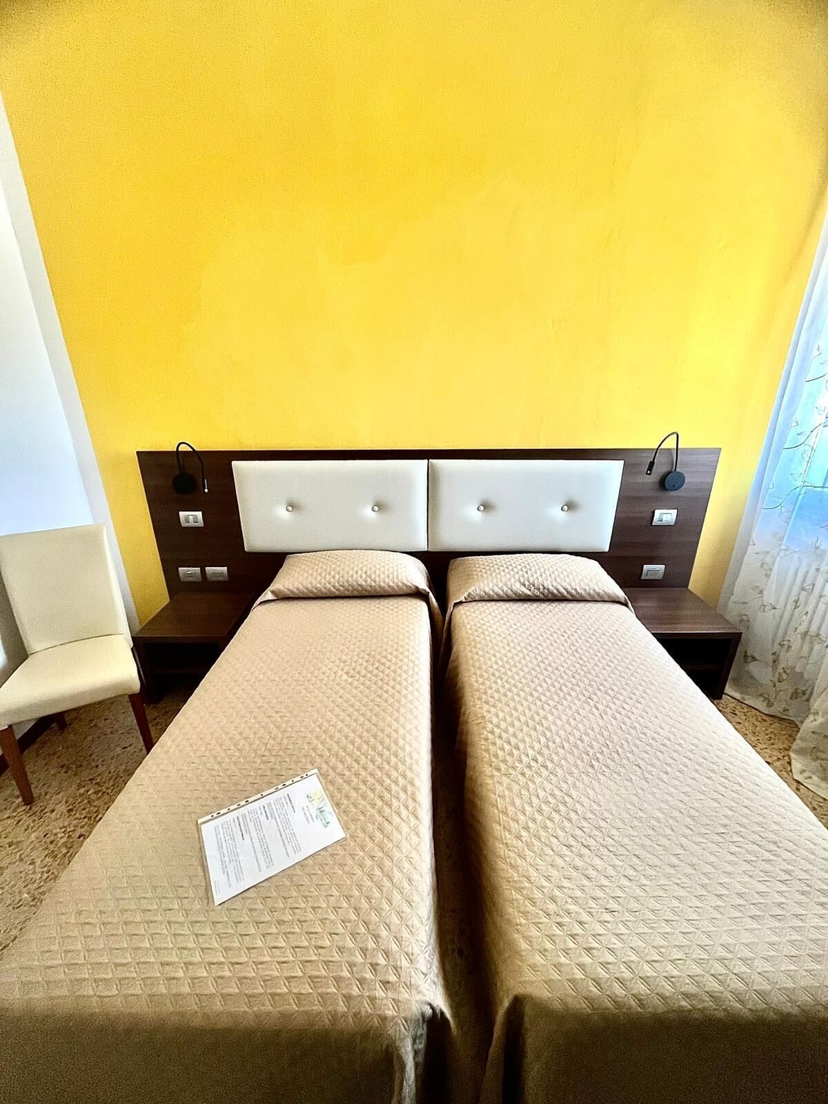 Camera d'albergo con due letti, una testiera, comodini, una sedia e una finestra con tende.
