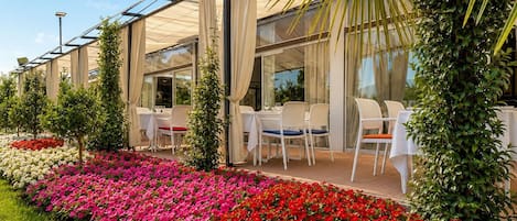 An outdoor dining area with tables and chairs, surrounded by flower beds with red and pink flowers, and a glass structure overhead.
