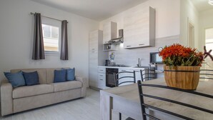 Una cocina moderna con armarios blancos, un comedor con mesa y sillas de madera, y una sala de estar con sofá y cojines.